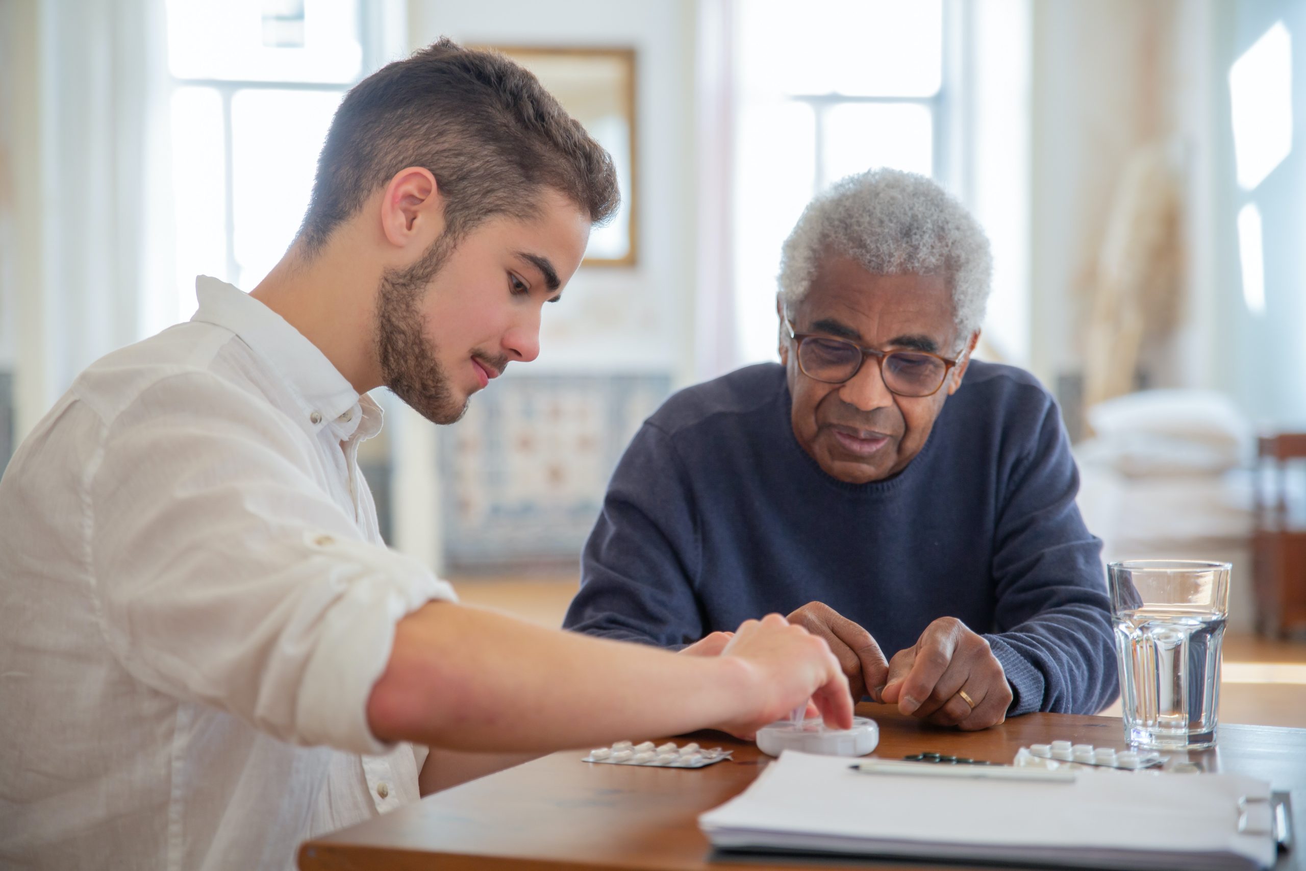 pexels-kampus-production-7551646 A man sorts medication at a table with an elderly man